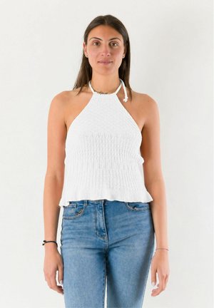 Donna che indossa un top bianco lavorato a maglia con collo a fascia e jeans blu, in piedi contro uno sfondo chiaro e uniforme con un'espressione neutra.