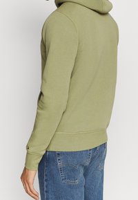 Person, der einen hellgrünen Hoodie und eine blaue Jeans trägt, von hinten und von der Seite gezeigt, mit entspanntem Arm an der Seite.