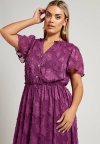 Lila klänning i lätt tyg med blomstermönster, korta puffärmar, v-ringning och knappfront, åtsittande i midjan.