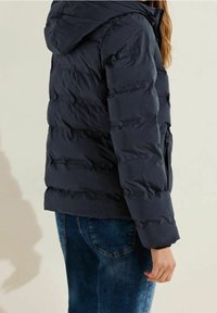 Granatowa kurtka puchowa z kapturem, z pikowanym wzorem, zapinana na zamek boczna kieszeń oraz miękka tekstura, zestawiona z niebieskimi dżinsami.