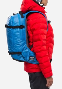 Blauer Rucksack mit strukturiertem Finish, verstellbaren Trägern und schwarzem Zubehör. Getragen über einer roten Daunenjacke, verfügt er über ein Marken-Patch.