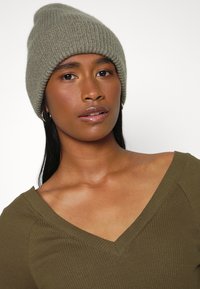 Gorro de malha canelada verde-azeitona com uma textura suave, usado com uma camisa verde-azeitona de decote em V profundo. Apresenta um ajuste justo e um design simples.
