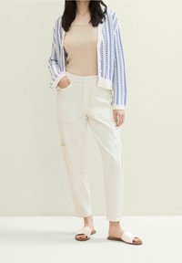 Witte cargobroek gecombineerd met een beige tanktop en een blauw-wit gestreepte gebreide cardigan. Beige slidesandalen met een platte zool.