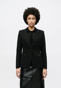 Μαύρο σακάκι ραμμένο κομψά από λείο ύφασμα, με εφαρμοστό σχέδιο, δύο μπροστινές τσέπες με χρυσές λεπτομέρειες και γιακά με χαραγή.