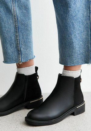 Stivali neri in pelle fino alla caviglia con zip laterale, dettagli in tessuto testurizzato, finitura dorata sul tacco, indossati con calze grigie lavorate a maglia e jeans blu.