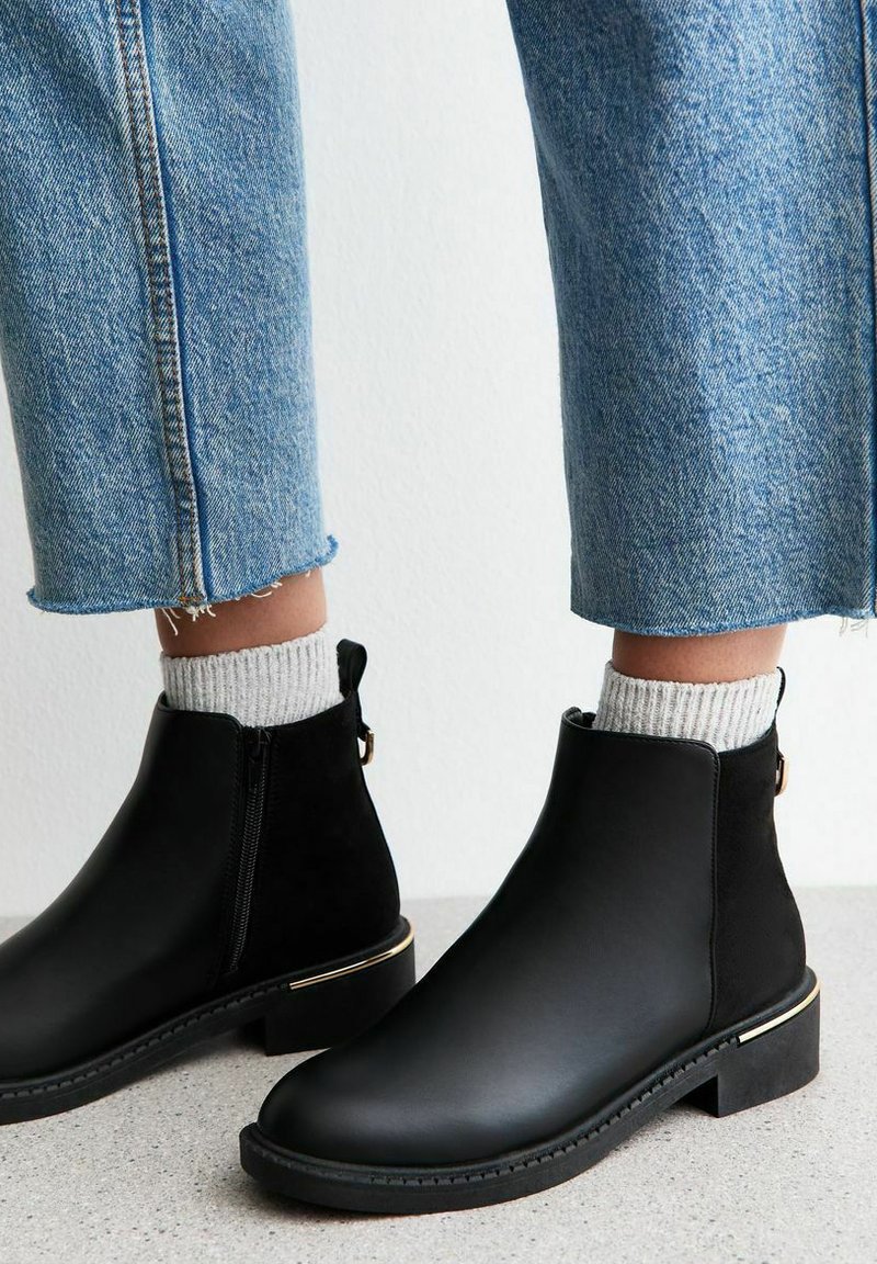 Botas de tobillo de cuero negro con cremallera lateral, acentos de tela texturizada, un embellecedor dorado en el tacón, y combinadas con calcetas de punto grises y denim azul.