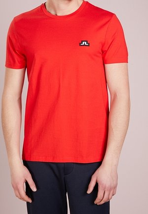 Mężczyzna w jaskrawoczerwonej koszulce z krótkim rękawem i okrągłym dekoltem, z małym czarno-białym logo na lewej piersi, w połączeniu z ciemnymi spodniami.