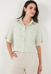 Camicia button-up verde chiaro a maniche corte con collo e due tasche frontali. Realizzata in un tessuto leggero con una finitura leggermente strutturata.
