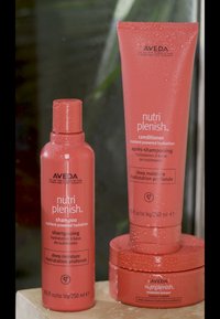 Aveda Nutri Plenish schampo och balsam i rosa flaskor med eleganta designer. Etiketterna lyfter fram återfuktande egenskaper. Vattendroppar på ytor.