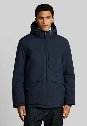 Mann trägt eine dunkelblaue Jacke mit Reißverschluss und Kapuze sowie Fronttaschen, steht vor einem einfarbigen hellgrauen Hintergrund.