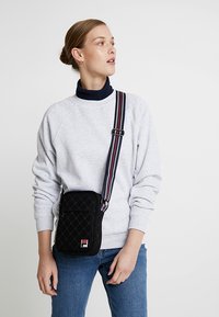 Borsa a tracolla nera trapuntata con tasca frontale con zip, dotata di una tracolla a righe e di un piccolo logo.