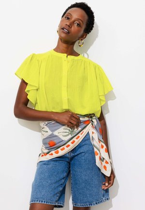 Femme portant un chemisier jaune vif à manches courtes, un short en jean bleu et une écharpe à motifs nouée autour de la taille, debout contre un mur blanc.