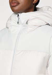 Leichte, weiße Steppjacke mit hohem Kragen, Reißverschluss und strukturiertem oberen Bereich. Mit dezenter Markenkennzeichnung auf der Brust und einer Kapuze.