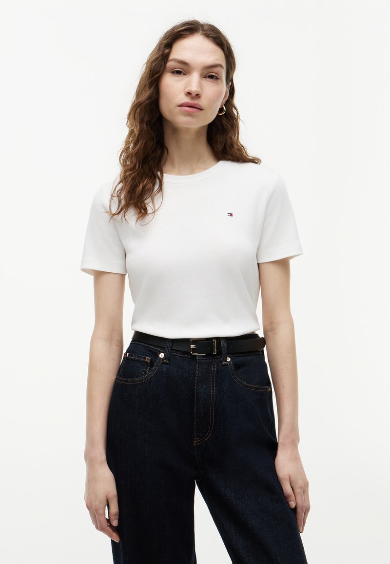 Donna con lunghi capelli mossi che indossa una maglietta bianca a maniche corte e jeans blu scuro con cintura nera su sfondo bianco uniforme.