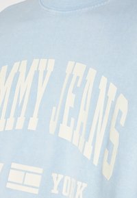 Sudadera azul claro con letras blancas en negrita que dicen "TOMMY JEANS" y "NEW YORK". La tela parece suave con un acabado texturizado.