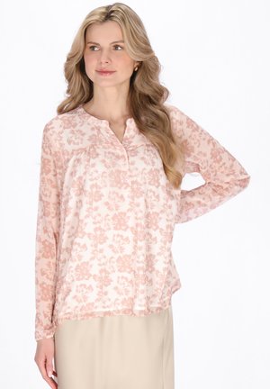 Kobieta z długimi blond włosami nosi jasnoróżową kwiecistą bluzkę z długim rękawem i beżową spódnicę, pozując z jedną ręką na biodrze.