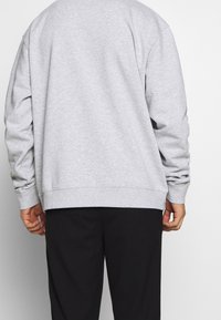 Lichtgrijze trui met lange mouwen, geribde boorden en een zoom, gecombineerd met zwarte broek. De stof lijkt zacht en gestructureerd.