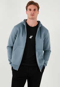 Sudadera con cremallera de color azul claro, con una textura suave, cordones en la capucha y dos bolsillos frontales, llevada sobre una camiseta negra.