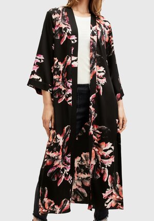 Nainen, joka pukeutunut mustaan, kukkakuviolliseen pitkähihaiseen kimonomalliseen avoimeen takkiin valkoisen paidan ja tummien farkkujen kanssa neutraalia taustaa vasten.