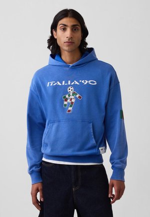 Tânăr purtând un hanorac albastru cu textul „ITALIA '90” și o grafică a unui jucător de fotbal în culorile steagului Italiei, asortat cu blugi închiși la culoare.