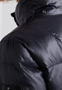 Nærbillede af skulder og øvre ryg på en sort polstret jakke med synlige sømme og en trykknap på kraven.