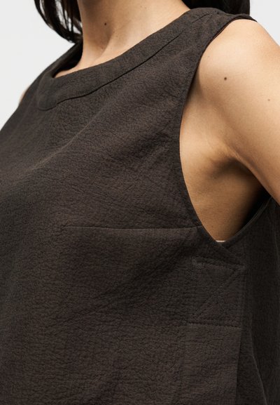 Καφέ αμάνικο τοπ από υφασμάτινη υφή, με στρογγυλή λαιμόκοψη, μικρό τσεπάκι στο στήθος και εμφανείς ραφές.