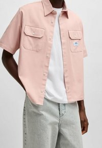 Homme portant une chemise à manches courtes rose clair avec des poches poitrine par-dessus un T-shirt blanc et un jean délavé, les mains dans les poches.