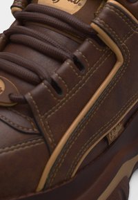 Κοντινή λήψη καφέ δερμάτινου αθλητικού παπουτσιού με χοντρά στρογγυλά κορδόνια, μπεζ ραφές και μικρή ετικέτα με την επωνυμία "Buffalo" στην πλευρική επιφάνεια.