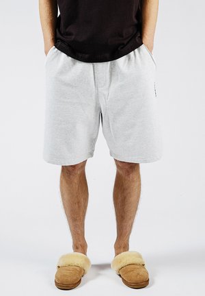 Man draagt lichtgrijze korte broek, zwart shirt en beige pantoffels met bontvoering, staand met handen in de zakken tegen een effen achtergrond.