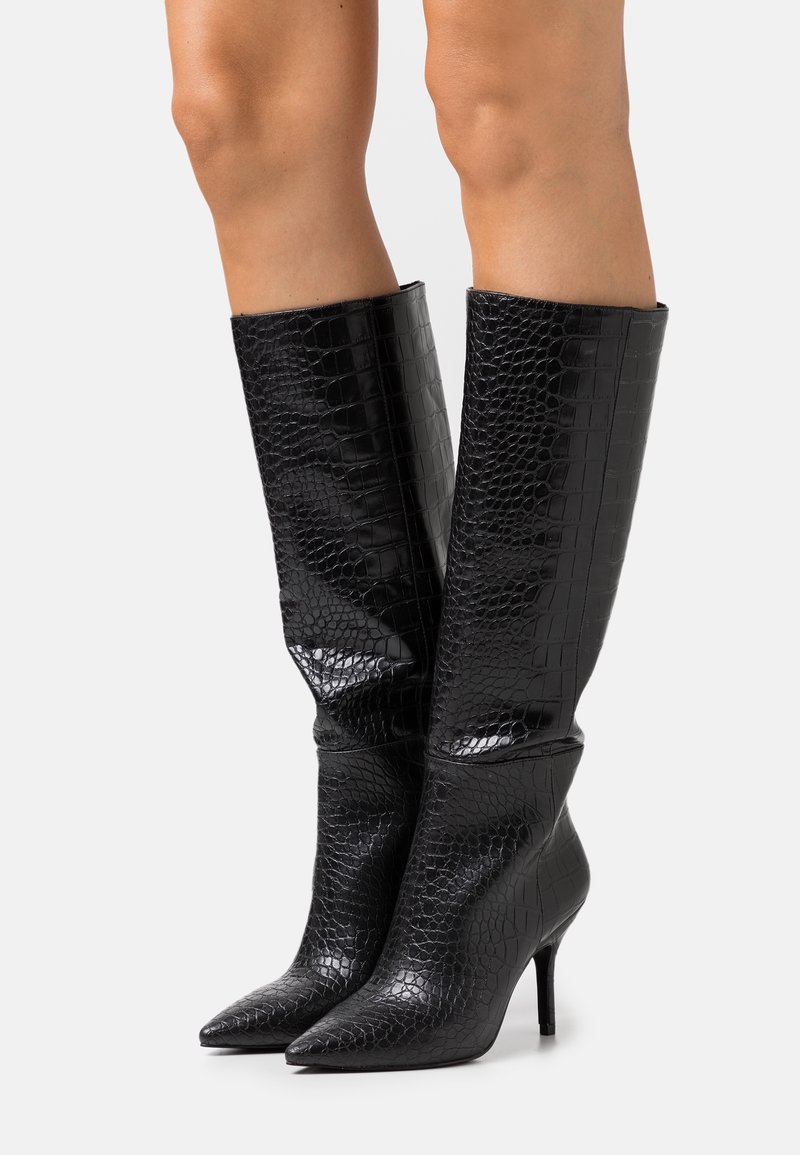 Bottes noires brillantes montant jusqu'aux genoux avec une texture crocodile, bouts pointus et talons hauts fins, portées sur des jambes nues sur un fond uni.