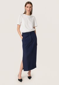 Weißes kurzärmliges Oberteil kombiniert mit einem marineblauen Midikleid. Der Rock hat seitliche Schlitze und eine taillierte Passform. Das Modell trägt schwarze spitze Absätze.
