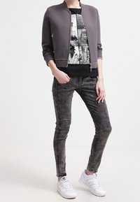 Chaqueta bomber gris sobre una camiseta gráfica negra, combinada con jeans skinny negros deslavados y zapatillas blancas. Atuendo urbano casual.