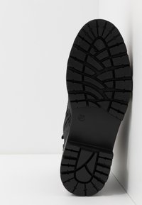 Semelle d'une botte noire avec un motif de bande de roulement profond et robuste, positionnée verticalement contre un mur blanc d'angle.