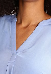 Ljusblå blus med v-ringning, tillverkad av mjukt tyg. Har subtila sömdetaljer vid kragen och en avslappnad passform.