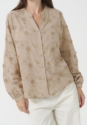 Beige langærmet bluse med blomsterbroderi og knapper, båret med hvide bukser, vist fra skuldre til hofter.