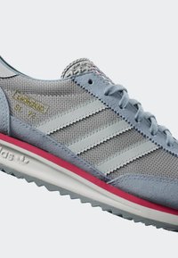Gray sneaker featuring a mesh and suede upper, three signature stripes, pink accent on the sole, and contrasting textured detailing.