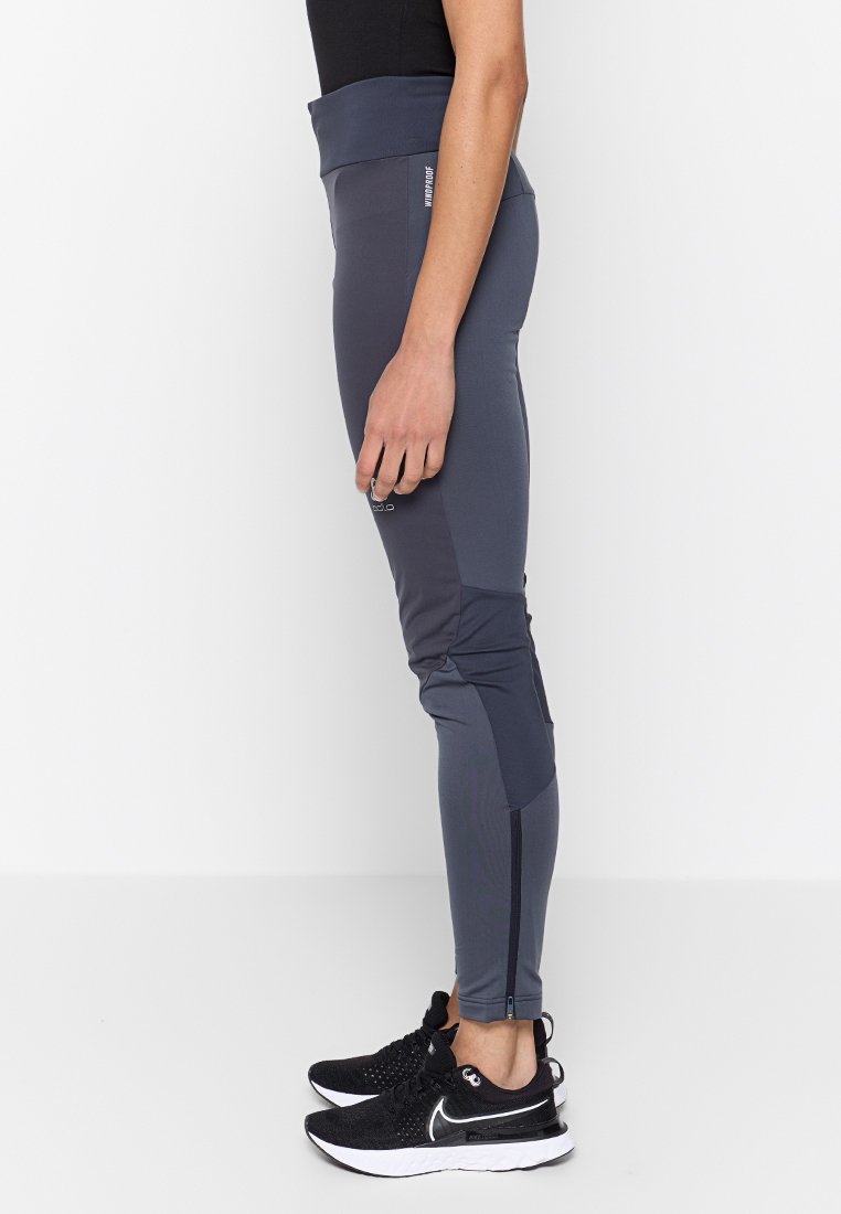 Graue Sportleggings mit hohem Bund, kontrastierenden Materialeinsätzen und Reißverschlüssen an den Knöcheln. Kombiniert mit schwarzen Laufschuhen.