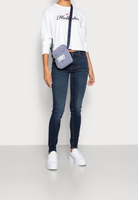 Blå denim skinny jeans i kombination med en vit långärmad skjorta med en logotyp. En liten blå axelväska b ärs över kroppen. Vita sneakers.