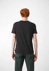 Schwarzes Baumwoll-T-Shirt mit rundem Ausschnitt und kurzen Ärmeln, mit lockerer Passform und Etikett an der linken Seitennaht. Getragen mit grünen Jeans aus Denim.