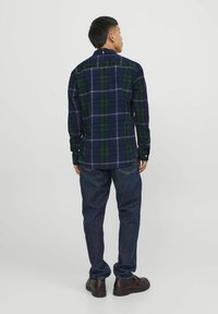 Camisa a cuadros en azul marino y verde con cuello abotonado, mangas largas y dobladillo curvado, combinada con jeans azul oscuro y zapatos marrones.