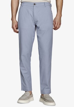 Mężczyzna ubrany w jasnoniebieskie spodnie o kroju slim-fit i beżowe buty wsuwane, stojący na białym tle.