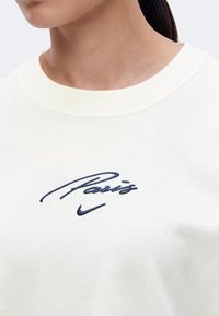 Gros plan d'une personne portant un t-shirt blanc à col rond avec « Paris » et le logo Nike Swoosh brodés en bleu marine sur la poitrine.