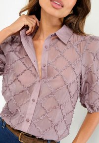 Gennemsigtig, mauve bluse med et struktureret diamantmønster og flæsedetaljer. Har en knappet front og puffede ærmer.