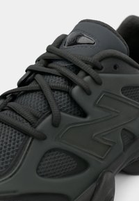 Gros plan d'une chaussure de sport noire avec un tissu en mesh, des lacets épais et un grand logo "N" en relief sur le côté.