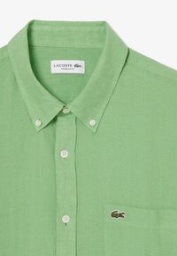 Camicetta verde in lino a maniche corte con colletto, dotata di bottoni bianchi e una tasca sul petto sinistro decorata con un piccolo logo di coccodrillo.
