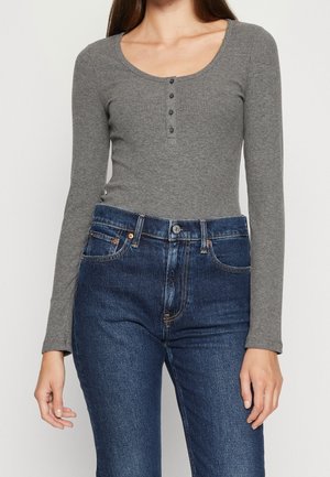 Vrouw draagt een aangesloten grijs geribd shirt met lange mouwen en knopen, gecombineerd met een donkere, hooggetailleerde spijkerbroek, staand tegen een witte achtergrond.