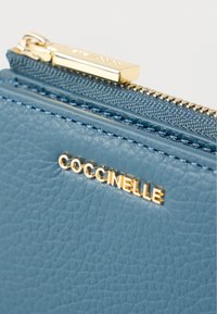 Sininen, kuvioitu nahkainen korttikotelo, jossa on kultaiset metalliosat, vetoketjusuljenta ja etupuolella kohokuvioitu brändinimi "COCCINELLE".