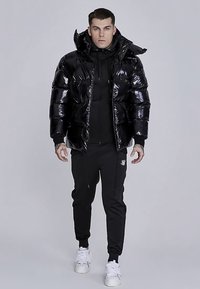 Skinnende sort dunjakke med hætte, lynlås foran og elastiske manchetter, lagdelt over en sort hættetrøje og kombineret med sorte træningsbukser og hvide sneakers.