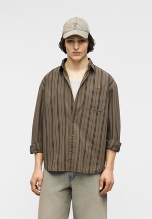 Fiatal férfi bézs sapkában, túlságosan nagy barna csíkos ingben, felhajtott ujjakkal, világosszürke farmerben, letisztult háttér előtt állva.