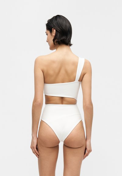 Γυναίκα με κοντά σκούρα μαλλιά, που φοράει λευκό μπικίνι τοπ με έναν ώμο και μπικίνι με ψηλή μέση, φωτογραφημένη από πίσω σε απλό φόντο.
